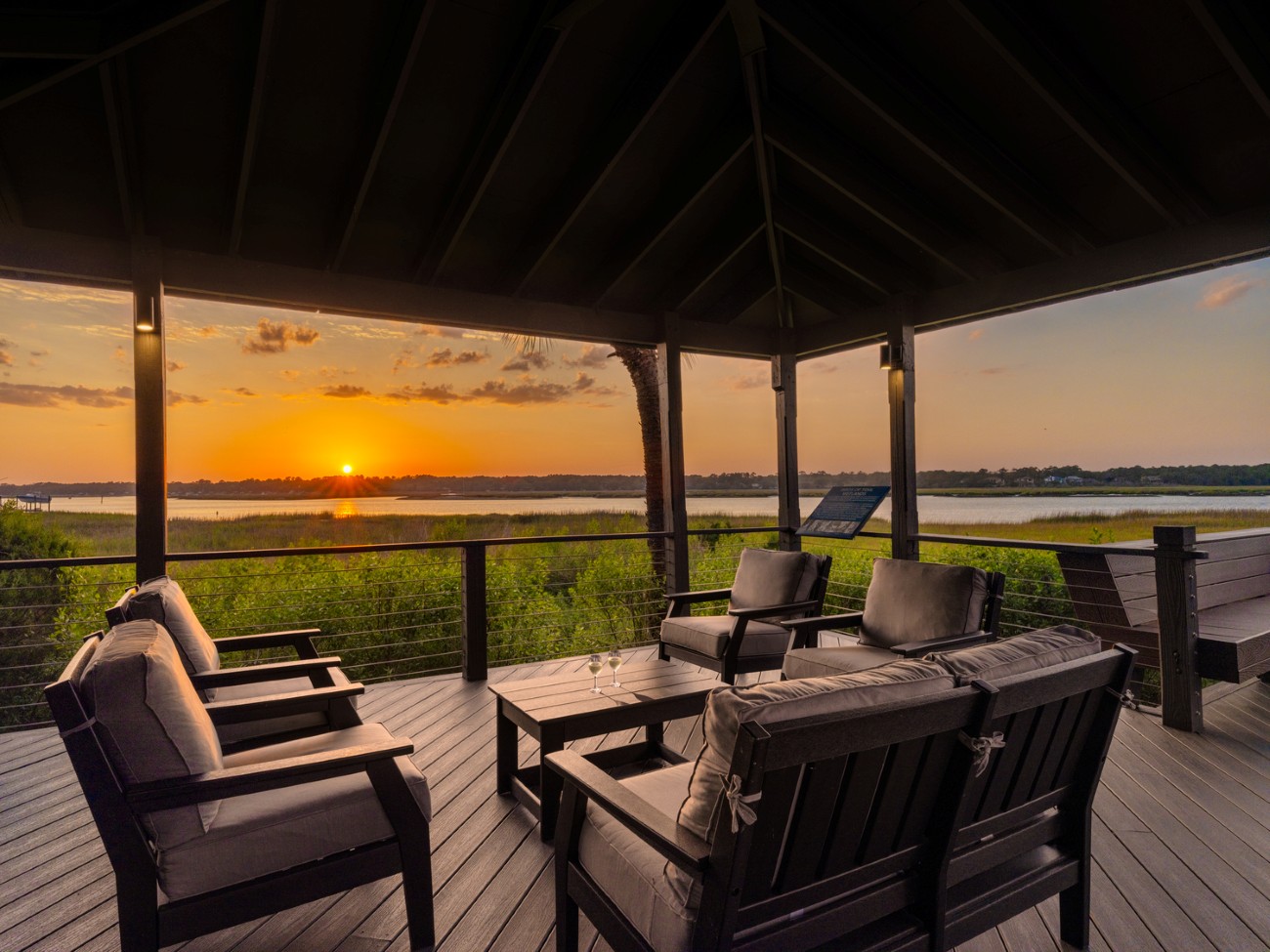 3.	Two glasses of wine sit on a table in between seating on a deck overlooking the marsh as the sun begins to set.