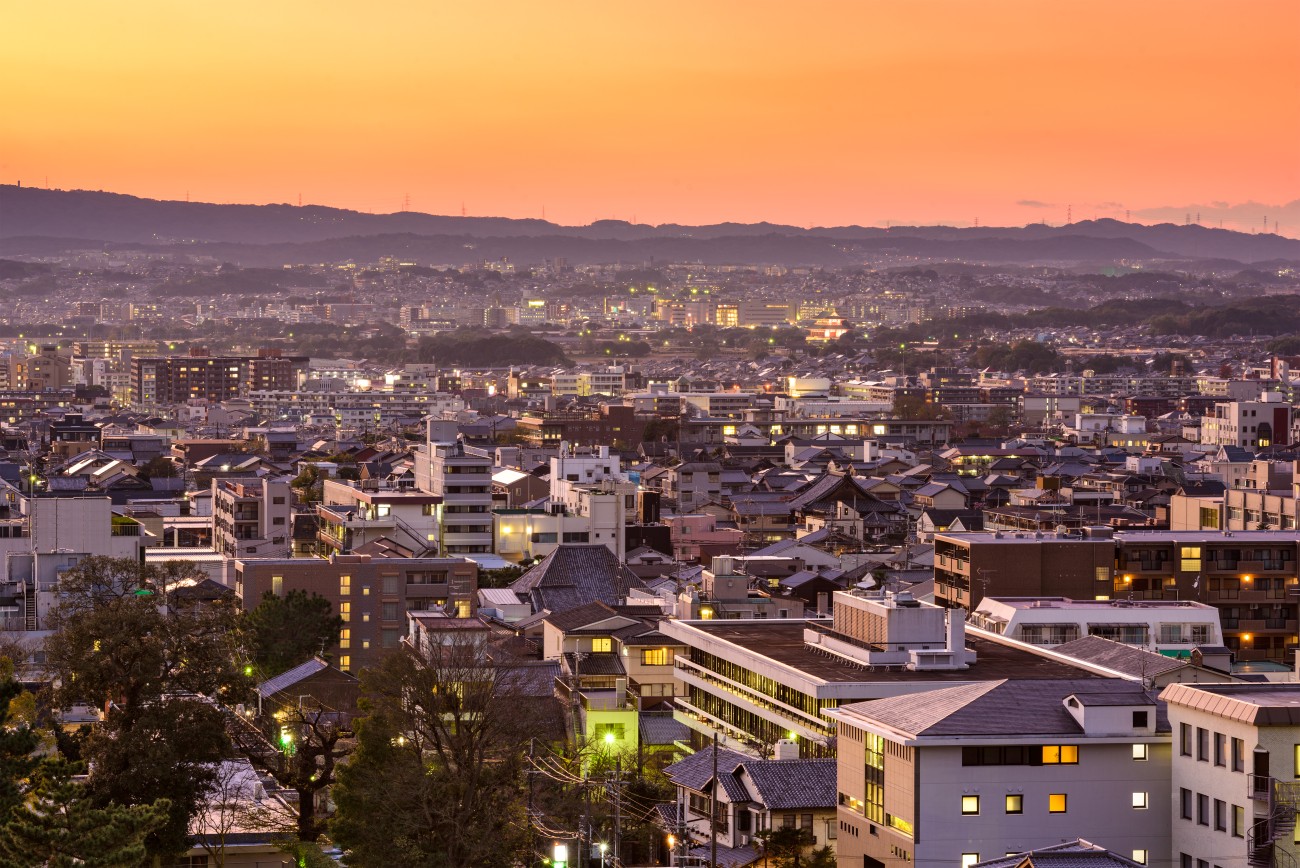 A city sits under a clear sky as the sun begins to set, and the lights begin to turn on.