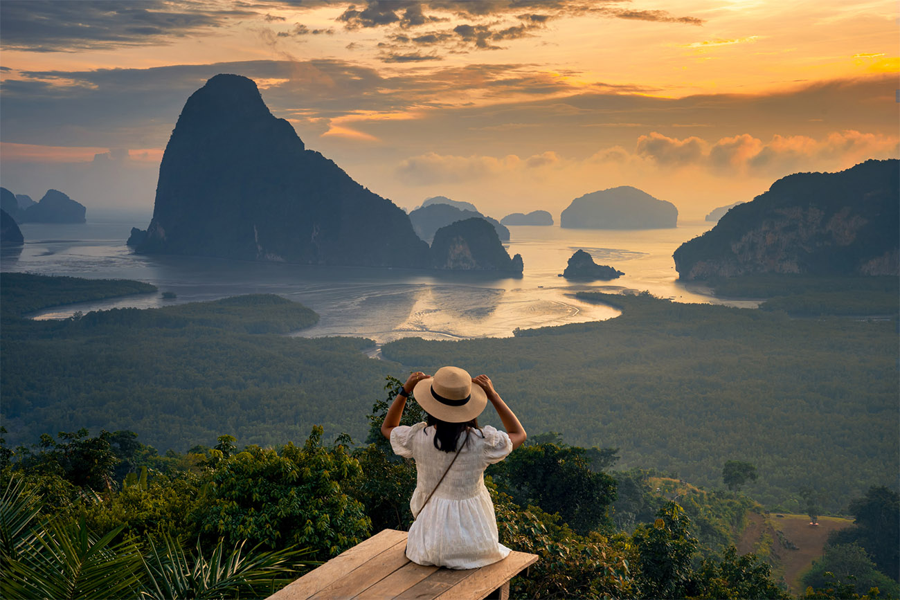 A person sits on the edge of a green hill overlooking a valley filled with trees and calm waters.