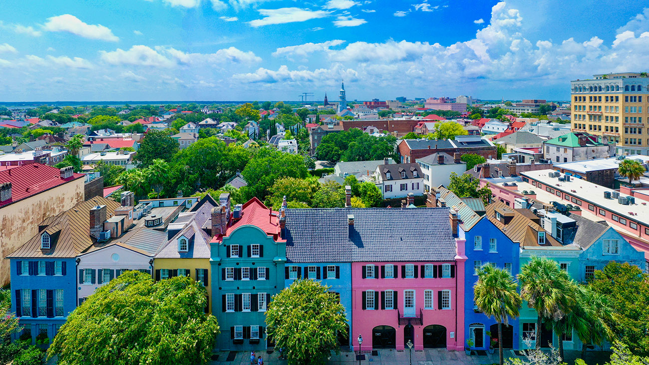 Aerial view view of Charleston, South Carolina, with colorful historic brick buildings, cobblestone pavement, and green trees lining the street.