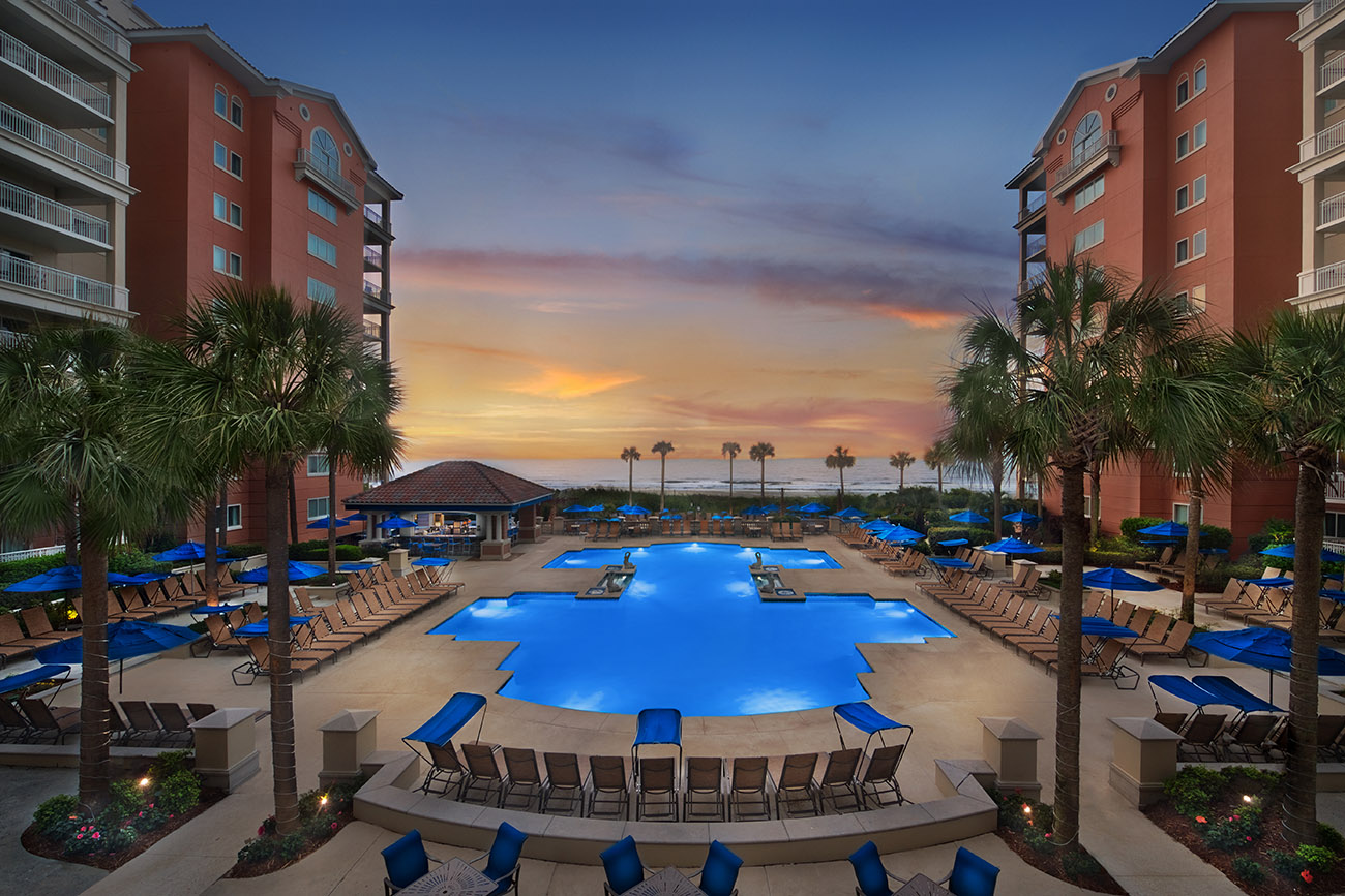 Aerial view of Marriott’s OceanWatch at Grande Dunes® with multiple pools, palm trees, and lounge chairs arranged around the pool at sunset.