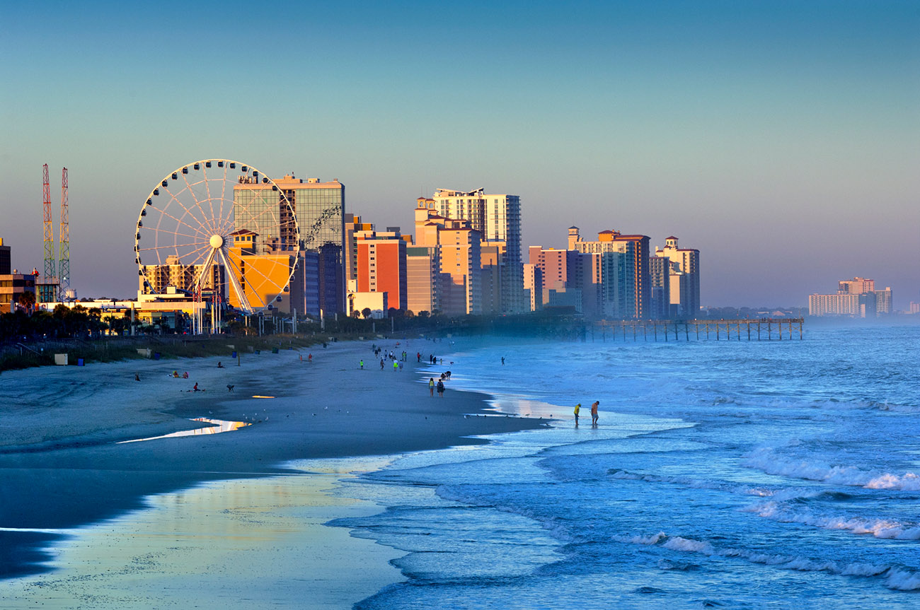 Aerial view of Myrtle Beach coastline with sandy beach, ocean waves, a Ferris wheel, and hotels lining the shore.