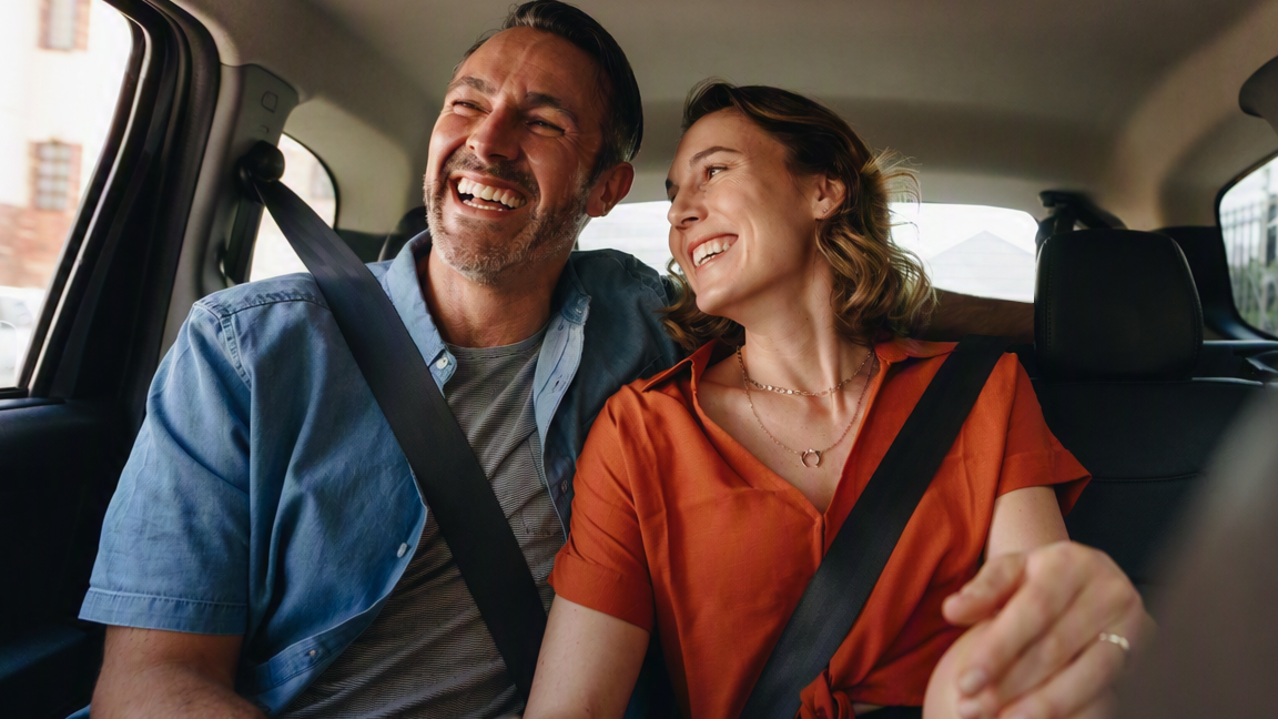 Two adults wearing seat belts sit together in the back seat of a car, with one holding a smartphone