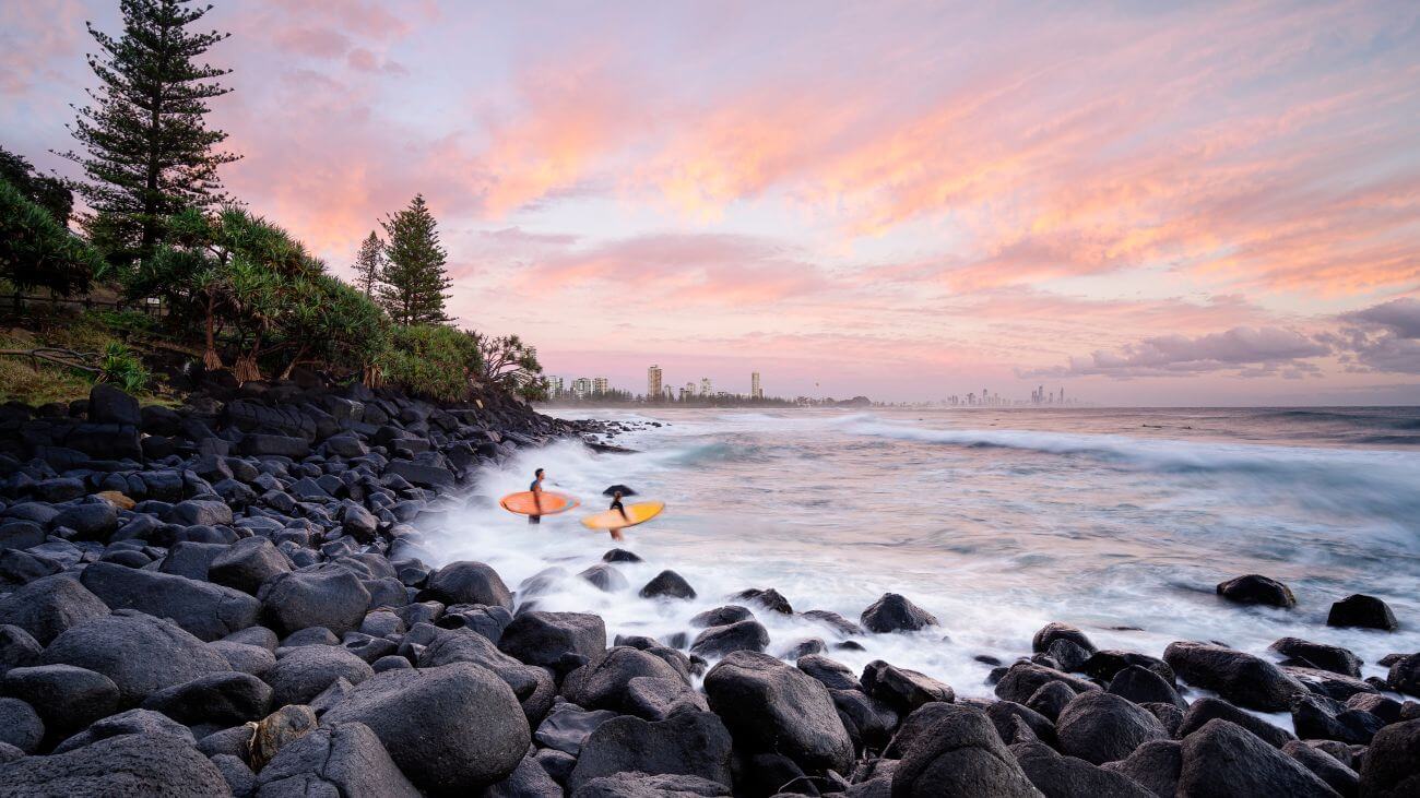 The sun begins to set behind a rocky coastline with waves hitting the rock.