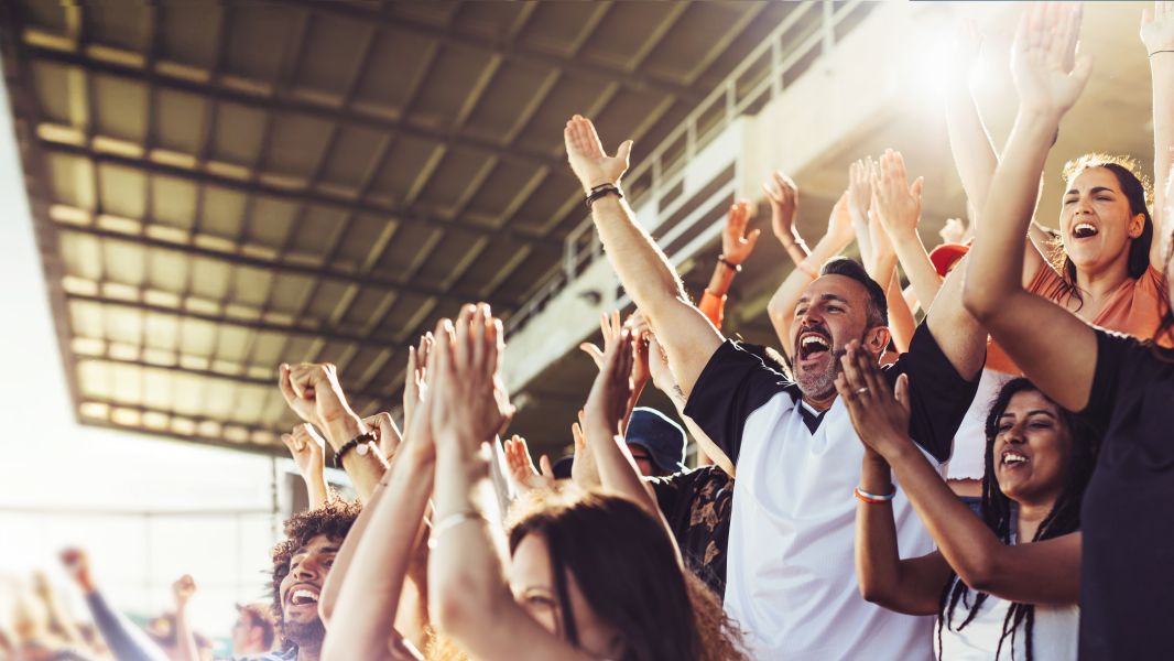 A group of fans loudly cheer with their hands in the air at a soccer game.