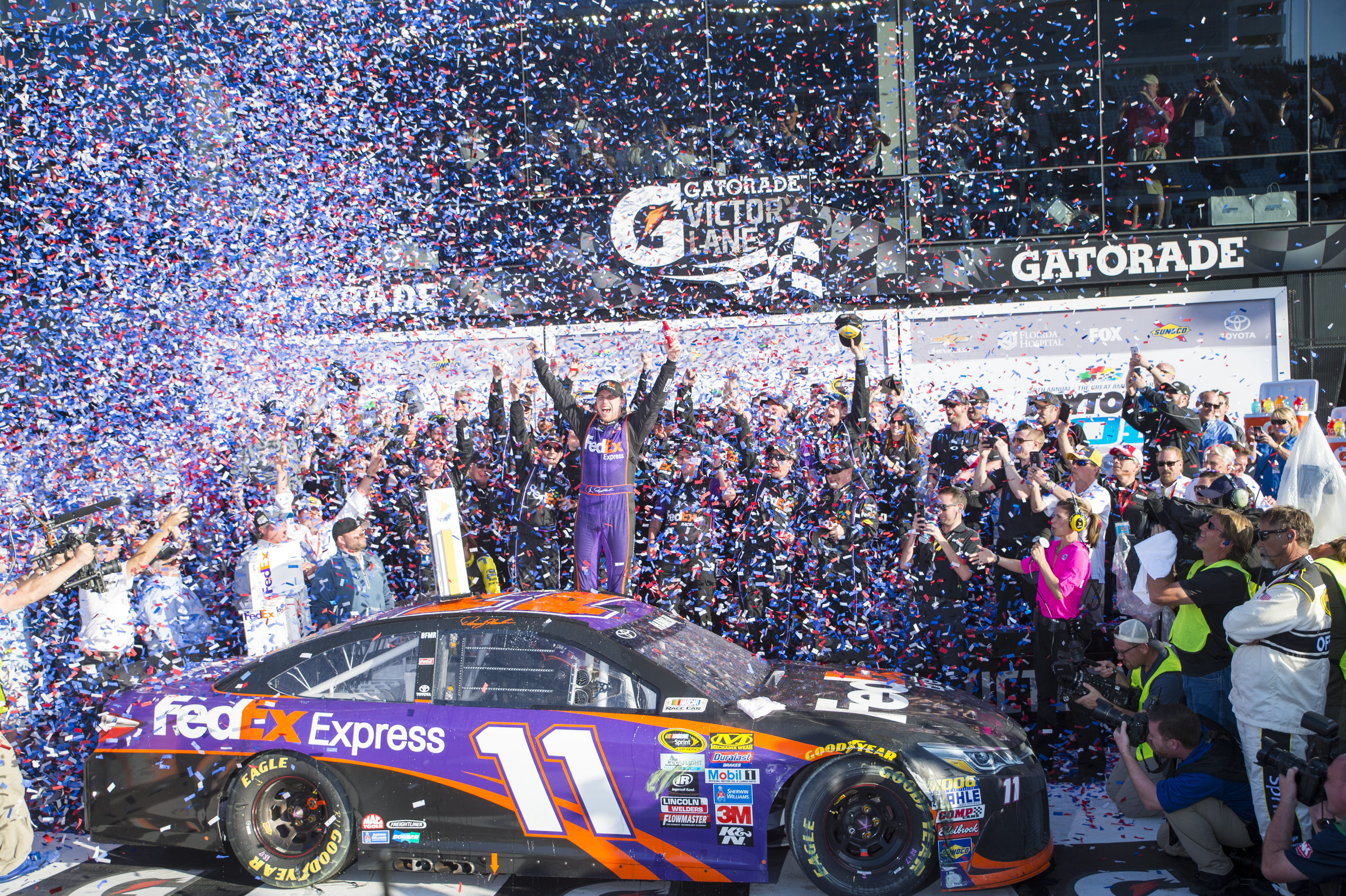 A winning NASCAR driver celebrates with his crew and fans in front of his car as confetti rains down.