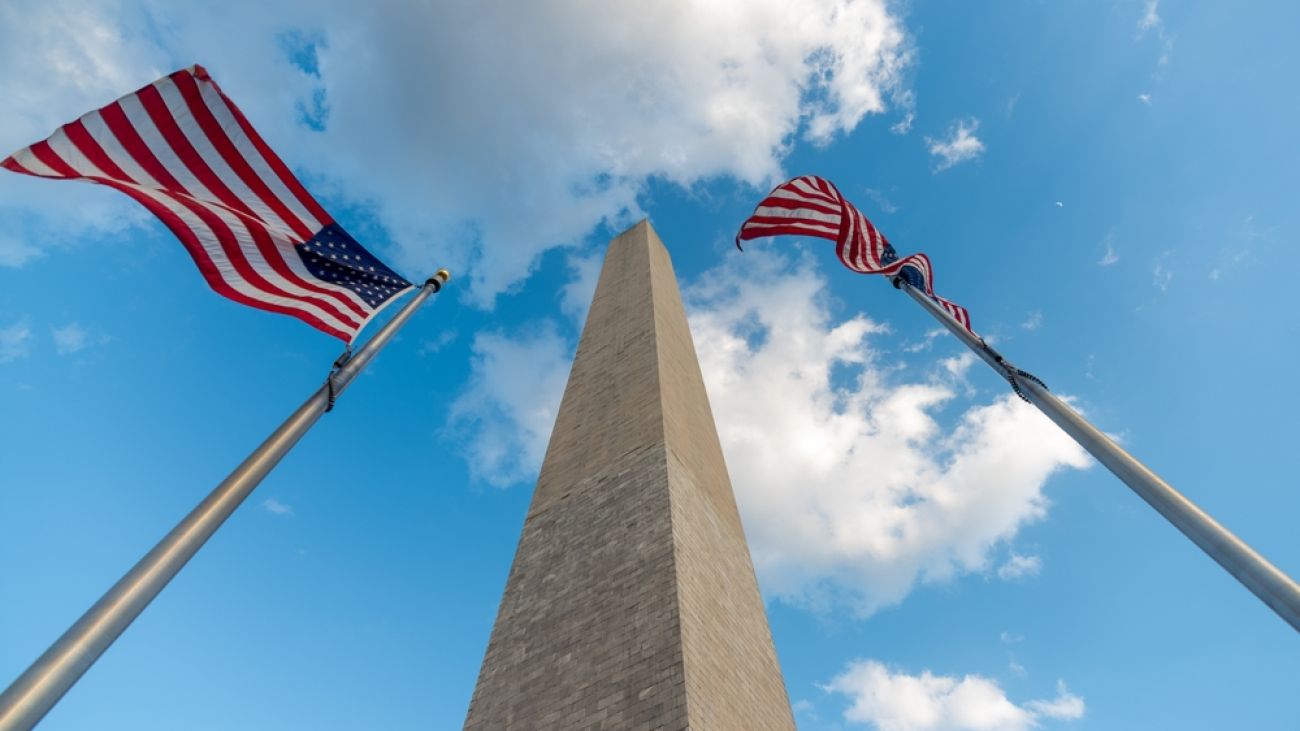 A low-angle view of the Washington Monument with two American flags waving in the sky.